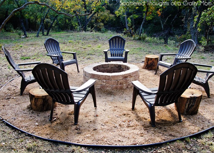 A circular brick fire pit with black plastic Adirondack chairs surrounding it in a backyard. 