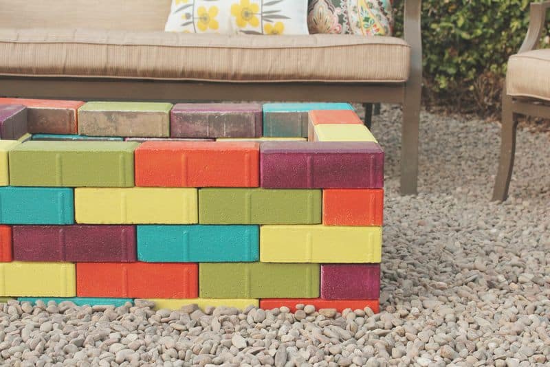 A colorful firepit made from green, yellow, teal, and purple bricks on a bed of gravel with an outdoor sofa.