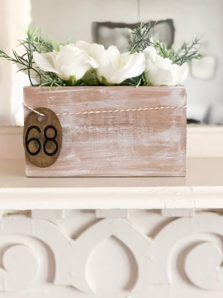 A whitewashed wood box with white flowers sits on a white antique side table.