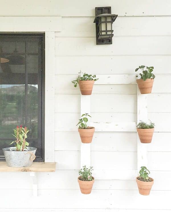 A white wooden wall hanging with clay pots and green plants inside on the side of a house.